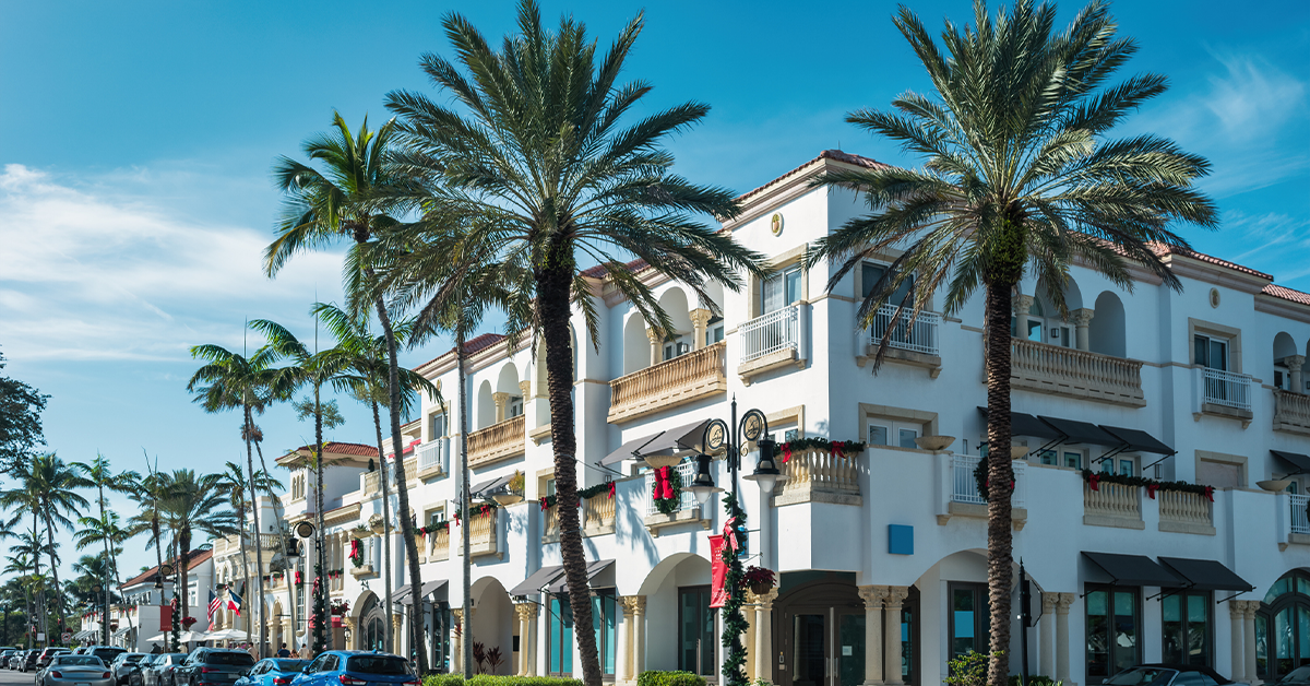 Palm-lined street with Mediterranean-style mixed-use building, storefronts, and parked cars