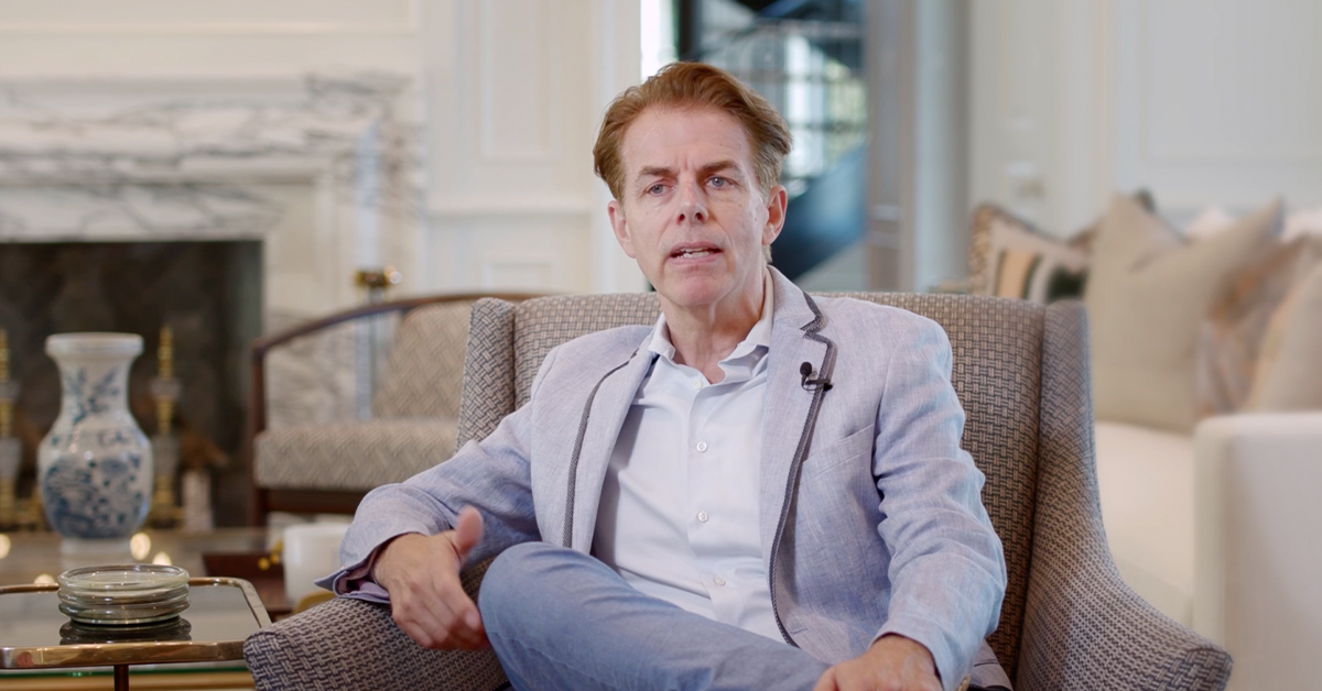 Man seated in armchair speaking during indoor interview.