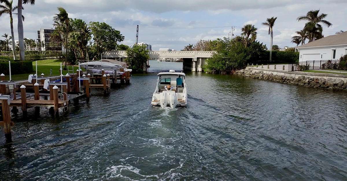 Motorboat moving down a canal between waterfront homes toward a low bridge under a cloudy sky.