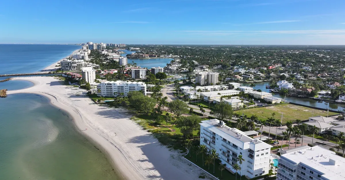 Aerial view of Naples, Florida's pristine white sand beach coastline with luxury waterfront condominiums and lush tropical landscaping under clear blue skies.